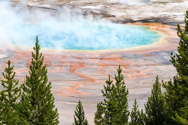 An aerial view of the Grand Prismatic Spring in Yellowstone National Park, showcasing its vibrant colors ranging from deep blues to vivid oranges and yellows. Steam rises from the spring, and the surrounding landscape is a mix of green forests and geothermal features.