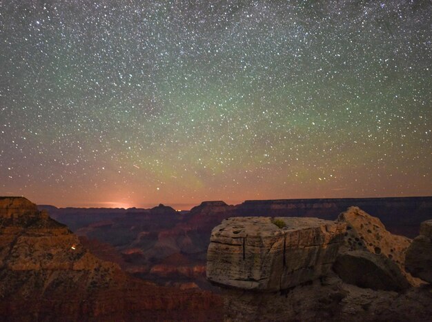 A wide-angle shot capturing the Milky Way galaxy arcing over the Grand Canyon at night, with the faint lights of the visitor center visible in the distance.