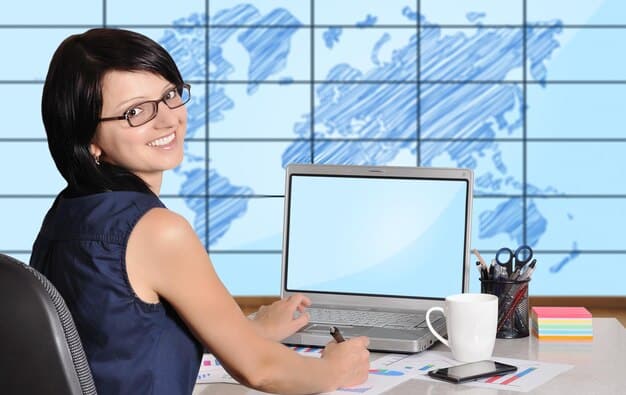 A friendly travel agent sitting at her desk, smiling and pointing at a computer screen displaying cruise deals. The office is bright and welcoming, with brochures and travel posters adorning the walls. A couple sits across from her, looking attentively at the screen.