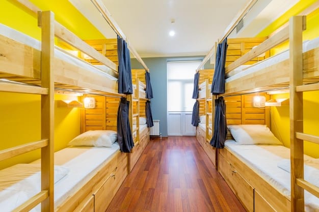 Photograph of a modern hostel dormitory with multiple bunk beds and individual lockers. Bright sunlight streams in through a large window, creating a welcoming atmosphere. Backpacks and travel essentials are neatly placed around the beds, suggesting a comfortable and organized space for student travelers.